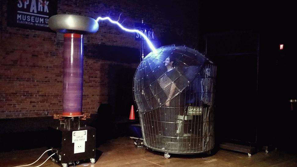 A man inside a Faraday cage being protected from a large arcing blue bolt of electricity being produced by a Tesla coil.